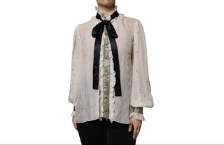A woman wearing a sheer white long-sleeve blouse with a black satin bow at the high neck, a ruffled front panel, and subtle gold dot embroidery, paired with black pants against a white background.