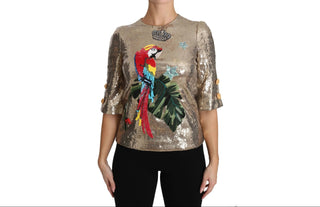 A woman wearing a metallic gold sequined short-sleeve top with a large colorful parrot and green leaf graphic on the front, paired with black pants, against a plain white background.