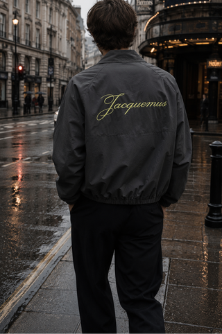 A man viewed from behind wearing a dark gray bomber jacket with 'Jacques' embroidered in gold script on the back, standing on a wet city street in London near the Theatre de Versailles.