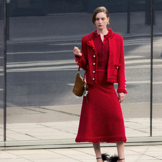 A woman stands in front of a glass storefront wearing a bright red coordinated outfit consisting of a cropped jacket, top, and midi skirt, with black ankle boots and a small brown handbag. Her reflection is visible in the glass to the left.