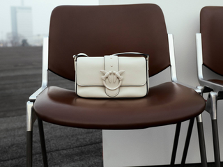 A cream-colored leather shoulder bag with a large horsebit clasp, displayed on a brown chair.