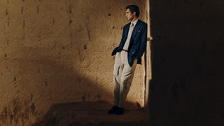 A man stands in a dimly lit, textured stone or plaster room wearing a dark navy blazer, cream dress shirt, cream trousers, and dark dress shoes.