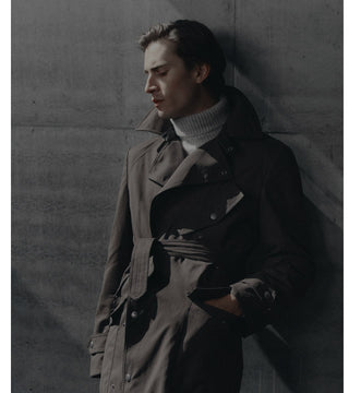 A man wearing a dark brown trench coat over a white turtleneck sweater, leaning against a textured gray concrete wall.
