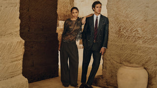 A man and a woman pose together in a rustic stone-walled setting. The woman wears a sheer black lace top and dark wide-leg trousers, while the man wears a dark double-breasted suit with a white shirt and a dark red tie.