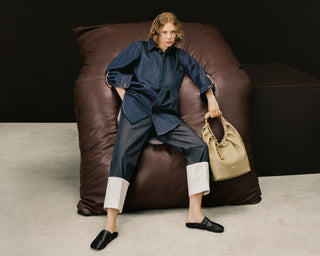 A model sits on a large brown beanbag chair in a studio setting, wearing a dark blue denim shirt and matching denim trousers with white cuff tabs, black loafers, and holding a beige drawstring handbag.