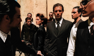 A group of men in dark suits and ties stand against a warm beige wall, with one man in the center wearing a pinstripe suit and tie. A woman in a dark blazer is visible beside him, and another man in the foreground is partially cropped on the left.