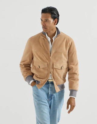 A man wearing a tan suede jacket with gray ribbed collar, cuffs, and hem, styled with a white collared shirt and light blue jeans against a light gray studio background.