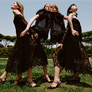 Three women pose outdoors in matching black lace midi dresses with black strappy heels, styled with black feathered vests, against a background of green trees and blue sky.