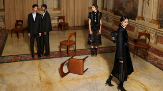 A fashion editorial scene in an ornate historic room with marble floors and wooden chairs. Three models are wearing dark tailored clothing: two men in black suits and a woman in a black lace dress, while another woman in a long black coat walks in the foreground.