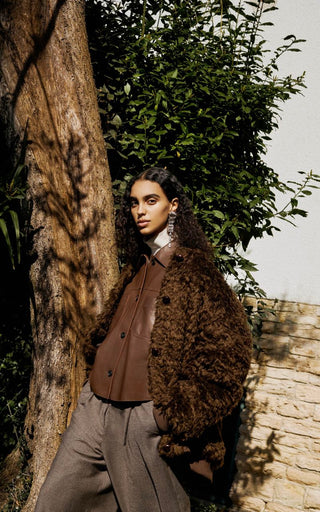 A woman wearing a brown textured jacket and gray trousers leans against a tree outdoors, with green foliage and a stone wall in the background.