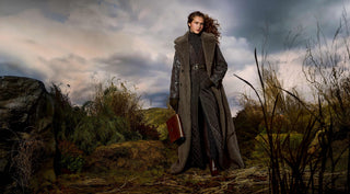 A woman stands outdoors in a dramatic, moody landscape under a cloudy sky, wearing a long dark overcoat over a dark patterned dress and holding a reddish-brown book or portfolio.