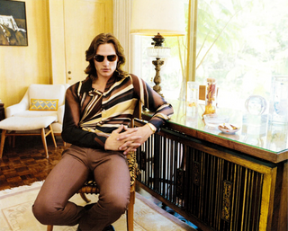 Man sitting in a chair wearing sunglasses in a room with a window and furniture.