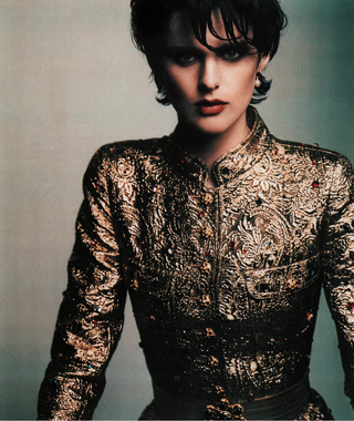 A model wearing a Chanel embellished gold and black patterned jacket, with the Chanel logo printed in black at the bottom on a white background.
