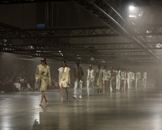A fashion runway show in a large industrial hall, with models walking down the catwalk in coordinated light-colored outfits under dramatic overhead lighting.
