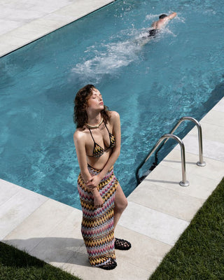 Woman in a colorful dress standing by a pool with another person swimming.
