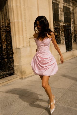A woman wearing a light pink off-the-shoulder mini dress and white pointed-toe heels walking on a stone sidewalk in front of a classical building.