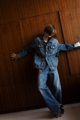 Person wearing a denim outfit standing against a wooden paneled wall.