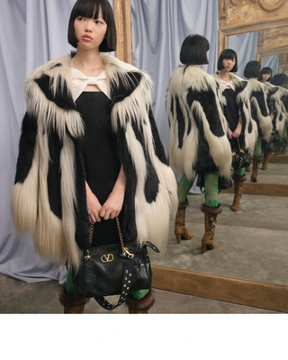 A model wearing a black and white patterned fur coat over a black dress, holding a black studded handbag with a gold chain strap. The photo is taken in a fashion showroom with a large mirror reflecting the model's back.