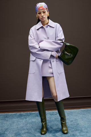 Person wearing a long light purple coat, green boots, and a colorful headscarf against a dark background.