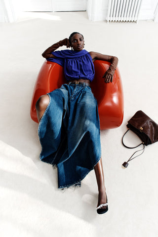 Woman sitting on a red chair wearing a blue top and jeans, with a brown handbag beside her on a white floor.