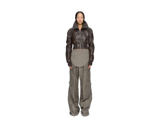 A model stands against a solid white background wearing a dark brown cropped leather jacket with a high collar, paired with a grey asymmetrical waist apron and wide-leg grey cargo pants.