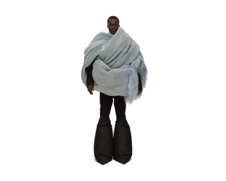 A model wearing a voluminous, light blue shaggy wrap or poncho draped over their shoulders, paired with black puffy boots, set against a solid white background.