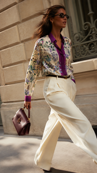 A woman walking outdoors in a stylish outfit featuring a long-sleeve floral print button-up shirt with a purple collar, cream wide-leg trousers, and a dark brown handbag.