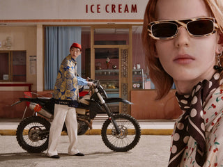 A fashion editorial scene featuring a model in a close-up on the right wearing tortoiseshell sunglasses and a patterned scarf, while another model on the left stands next to a dirt bike in front of an ice cream shop. The image has a strong Gucci aesthetic with colorful patterns and editorial styling.
