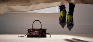 A brown leather handbag sits on a light floor beneath a bed, with a person's legs and neon green-and-black shoes dangling from the bed frame above.