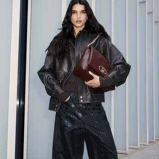 Person wearing a black leather jacket and holding a brown leather bag in an indoor setting.