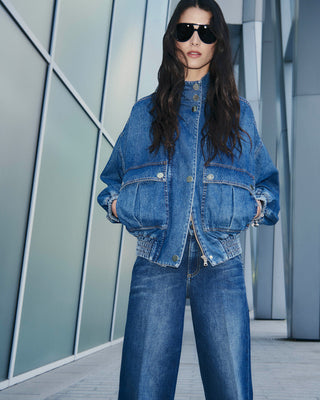 Person wearing a blue denim jacket and jeans standing in front of a modern architectural background.