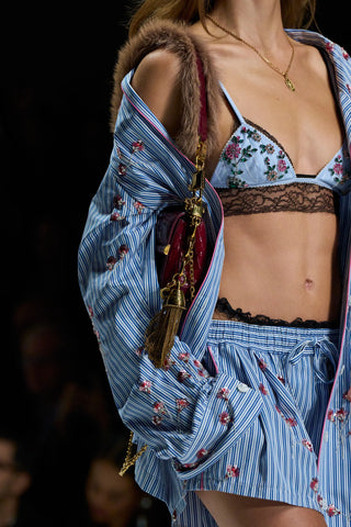 A close-up fashion runway shot of a model wearing a blue and white striped lingerie set with floral embroidery, a matching striped open jacket with faux-fur trim, and carrying a dark red tassel shoulder bag with gold hardware.