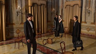 Four models in formal black clothing pose in an ornate, vintage-style room with gold drapes, marble floors, and wooden chairs.