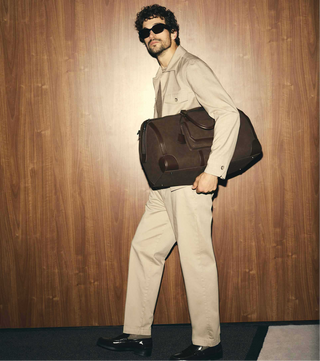 Man in beige jacket and sunglasses sitting on a leather chair, then standing with a brown bag.