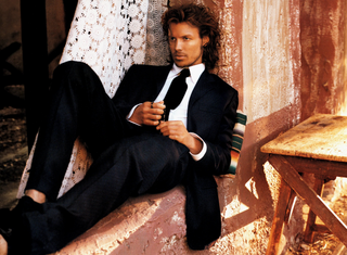 Man in a black suit sitting on a wooden stool with a textured wall and wooden floor in the background.