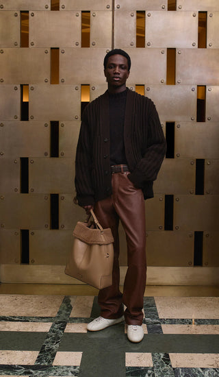 Man in dark sweater and brown pants holding a beige bag against a textured wall.
