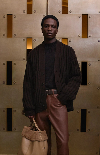 Man in a dark jacket and brown pants holding a tan bag against a textured wall.