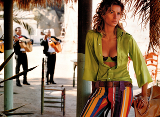 Woman in a green shirt and colorful pants standing under a palm tree with musicians in the background.